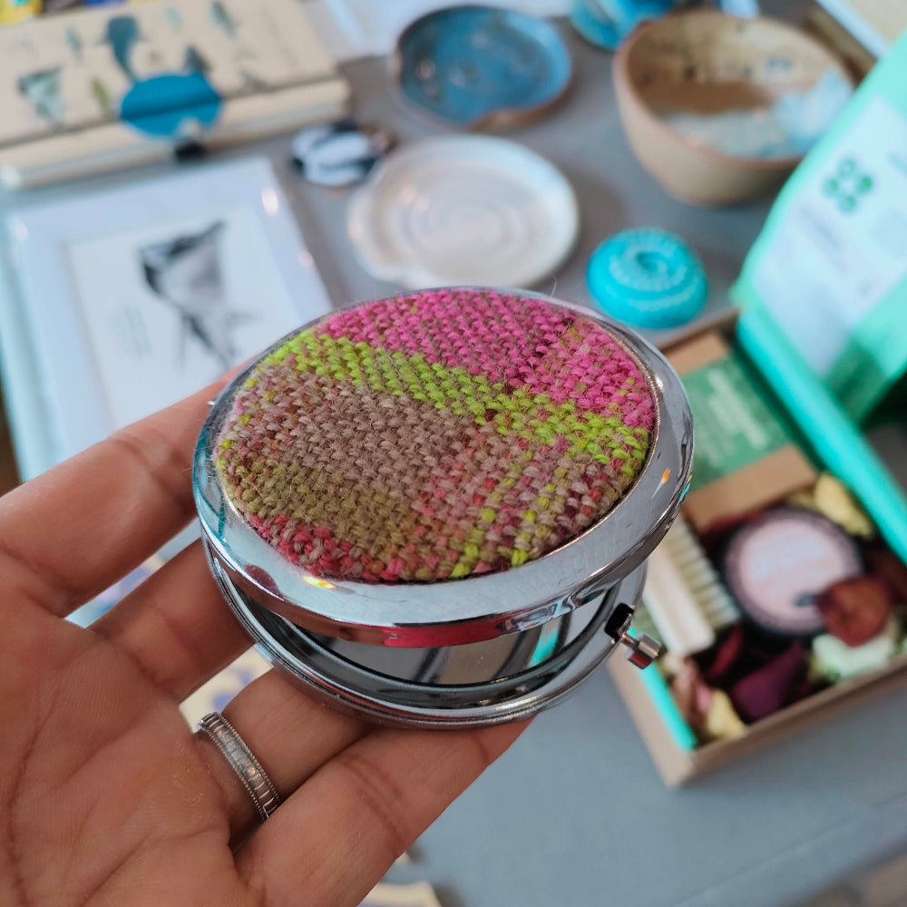 Hand holding a round, textured pocket mirror with a colourful top against a blurred background