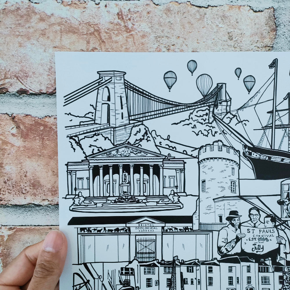 A hand holds a black and white illustrated art print depicting the Clifton Suspension Bridge, St Paul's Cathedral, and a sailing ship against a rustic brick wall background.