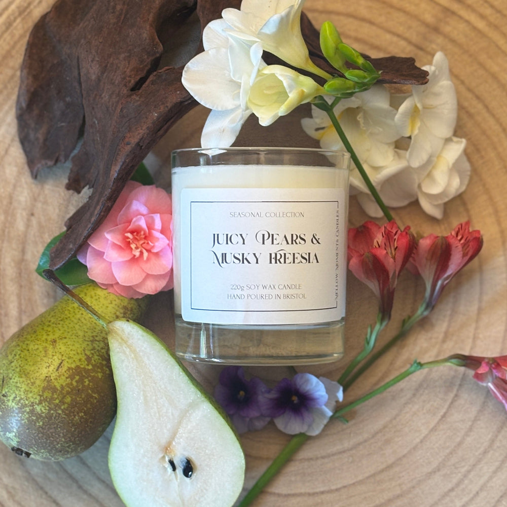 Candle labeled 'Juicy Pears & Musky Freesia' surrounded by pears and flowers on a wooden surface